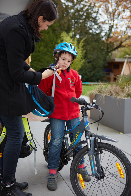 Mutter reicht einem Kind auf dem Fahrrad den Schulranzen an