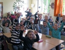 Blick in ein Klassenzimmer mit lachenden Schülerinnen und Schülern, die Ihre Arme in die Luft strecken.