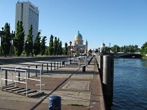 Ein breiter Gehweg führt an einem Kanal entlanf auf ein Hochhaus und eine alte Kirche zu.